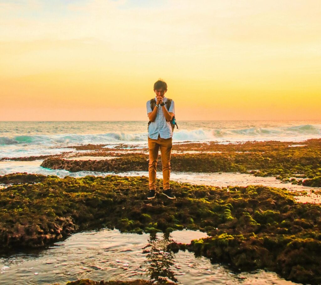 A man stands on mossy rocks by the ocean, praying during a vibrant sunset, creating a serene and introspective scene.